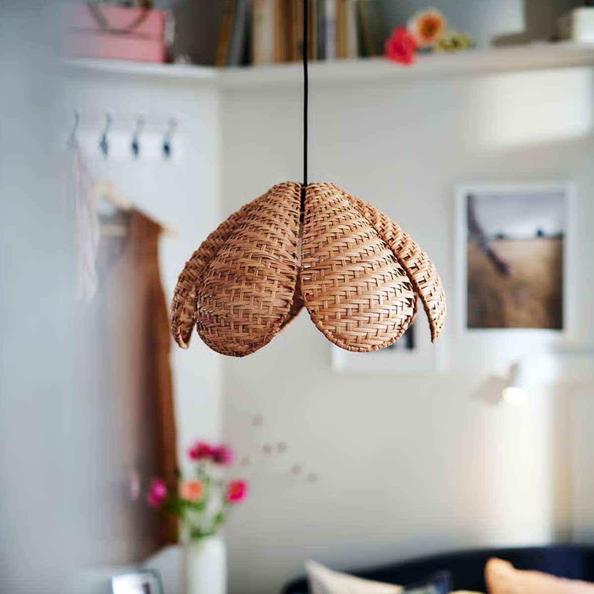 Lampada a sospensione in rattan intrecciato SANDKORN di IKEA con paralume a petali, appesa in una camera dallo stile caldo e naturale, dettagli di fiori e quadri sullo sfondo.