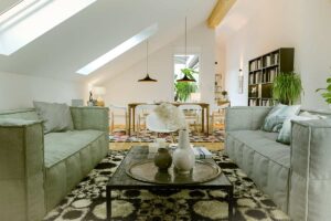 Soggiorno di mansarda moderna con soffitto spiovente, arredi bassi e pareti bianche. Due divani grigi e un tavolino centrale creano un’area living ariosa; sullo sfondo zona pranzo luminosa, piante verdi e libreria bassa, con luce naturale proveniente da finestre a tetto.