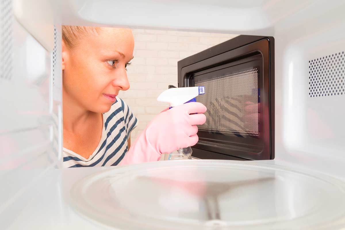 Donna con guanti rosa pulisce l’interno di un forno a microonde utilizzando uno spray, vista dall’interno dell’elettrodomestico.