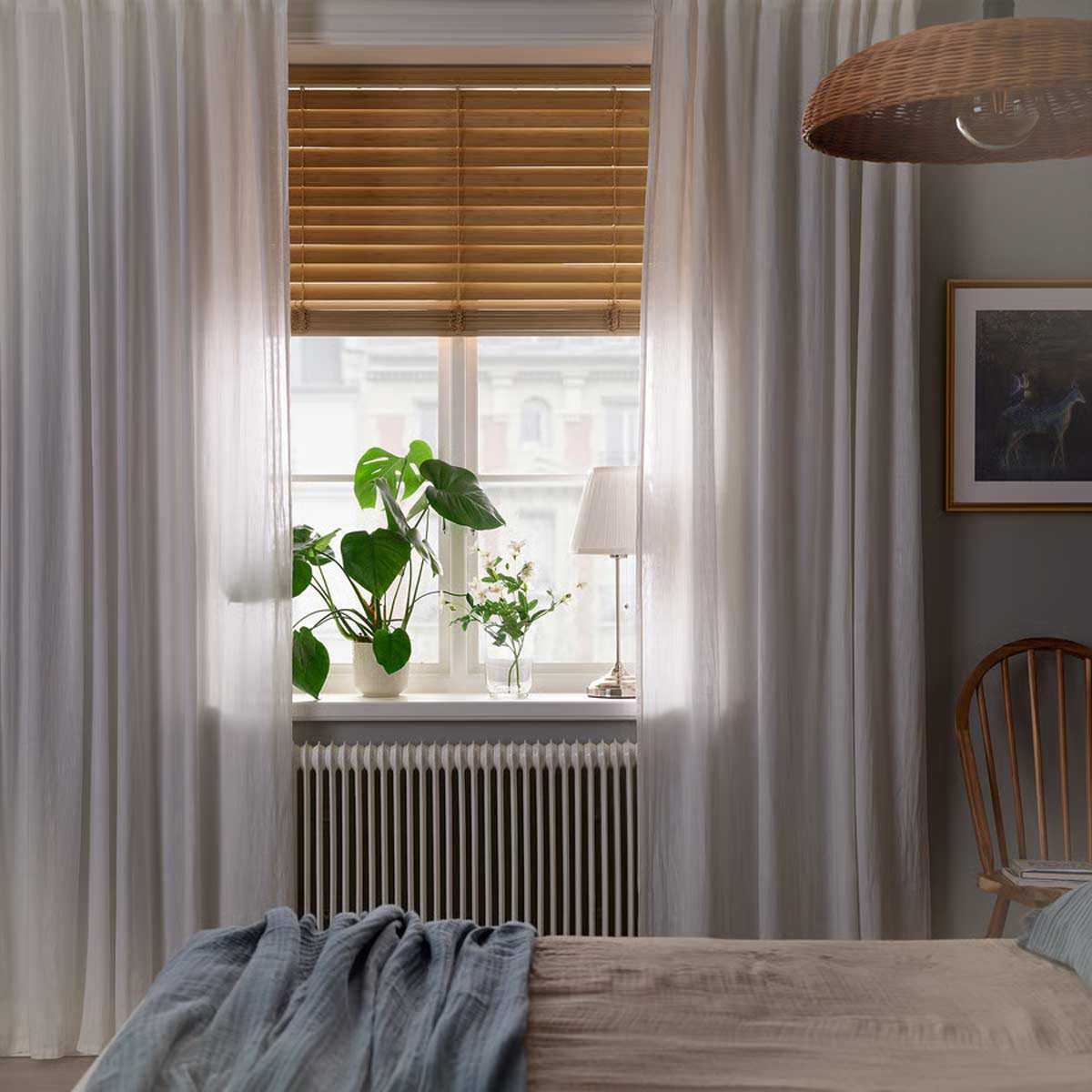 Camera da letto con veneziana in bambù naturale VECKLARFLY di IKEA, tende bianche leggere, piante verdi sul davanzale e luce filtrata per un ambiente rilassato e naturale.