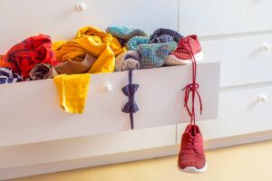 Cassetto di armadio bianco aperto con vestiti colorati e una scarpa rossa che pende fuori, segno di disordine nell’armadio dei bambini.