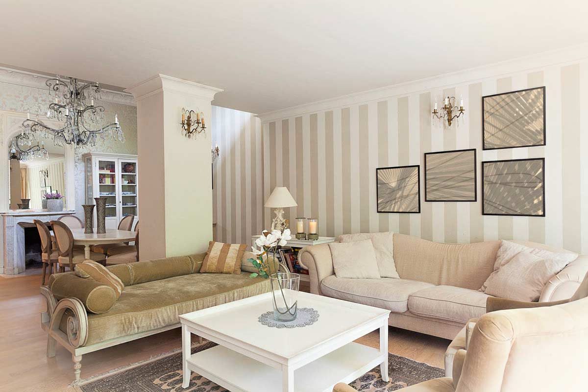 Salotto elegante con pareti tortora a righe verticali, divani chiari e tavolino bianco, dettagli classici e tocchi dorati. Sullo sfondo, sala da pranzo con lampadario in cristallo e arredi tradizionali.