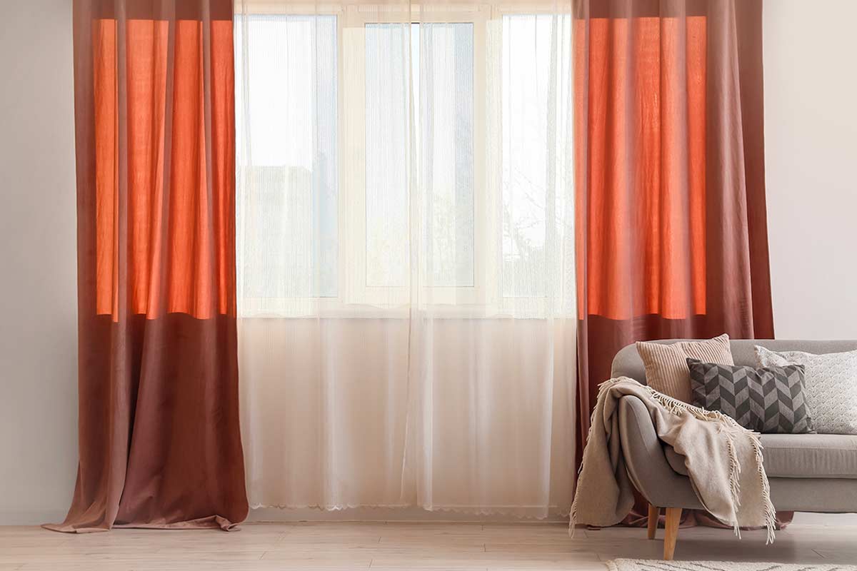 Tenda leggera color avorio davanti a una finestra ampia, affiancata da tende scure color terracotta, con divano grigio chiaro e cuscini in tessuti naturali. La luce filtra delicatamente nell’ambiente, creando un’atmosfera luminosa e accogliente.