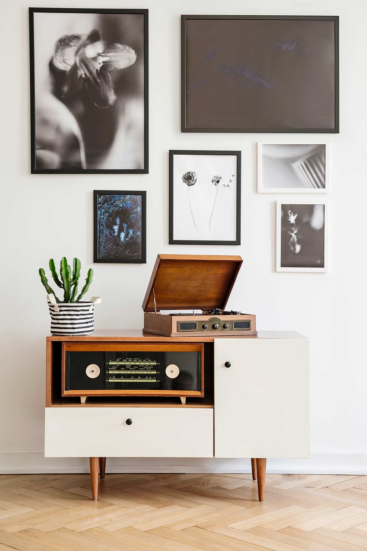 Credenza vintage anni ’60 con radio d’epoca e giradischi, inserita in una stanza moderna dal design essenziale. Parete decorata con quadri in bianco e nero e pianta grassa in vaso a righe. Esempio di armonia tra elementi retrò e arredo contemporaneo.