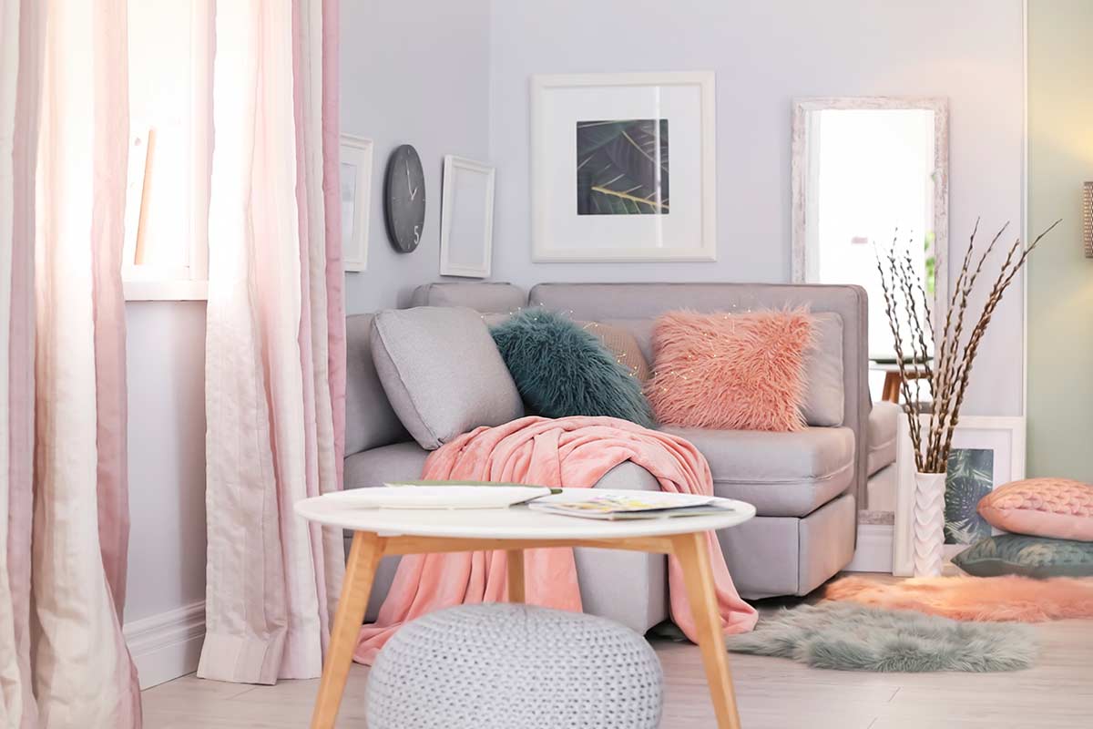 Soggiorno contemporaneo con divano grigio chiaro, cuscini e plaid nei toni polverosi del rosa cipria e verde salvia, tavolino rotondo bianco, tende rosa desaturato e dettagli di design che richiamano la palette anni '60.
