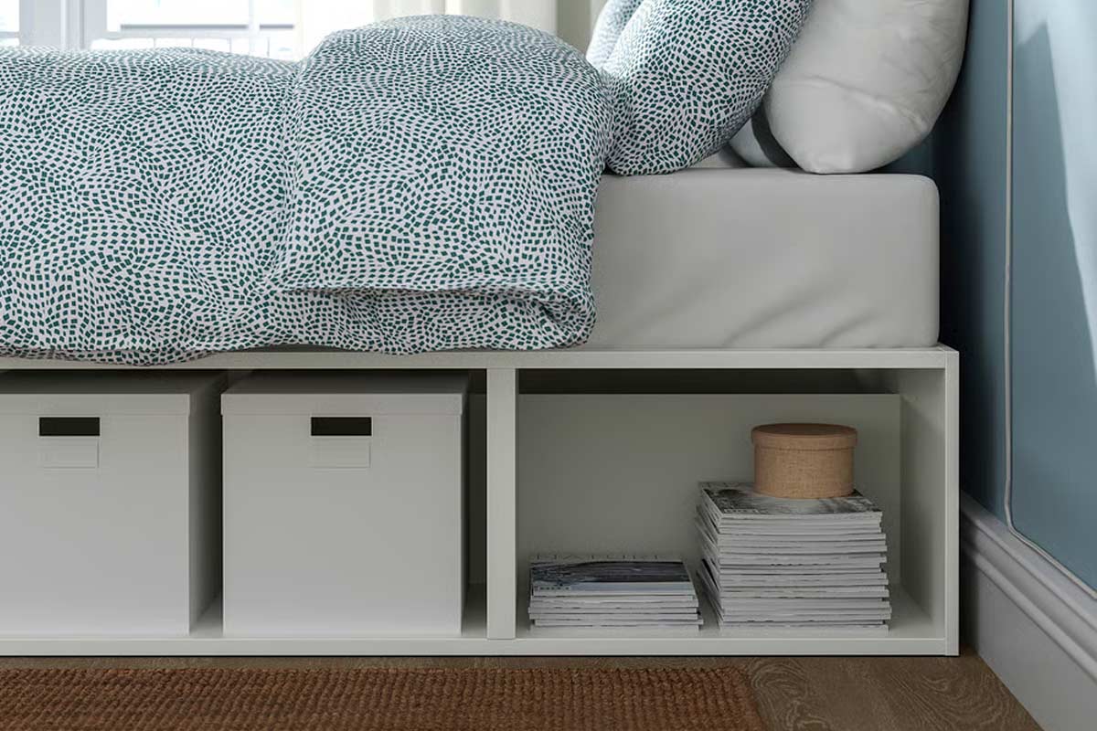 Dettaglio del letto contenitore IKEA PLATSA: vano laterale con scatole bianche, riviste ordinate e scatola tonda in tessuto, in una camera dalle pareti azzurre e luce naturale.