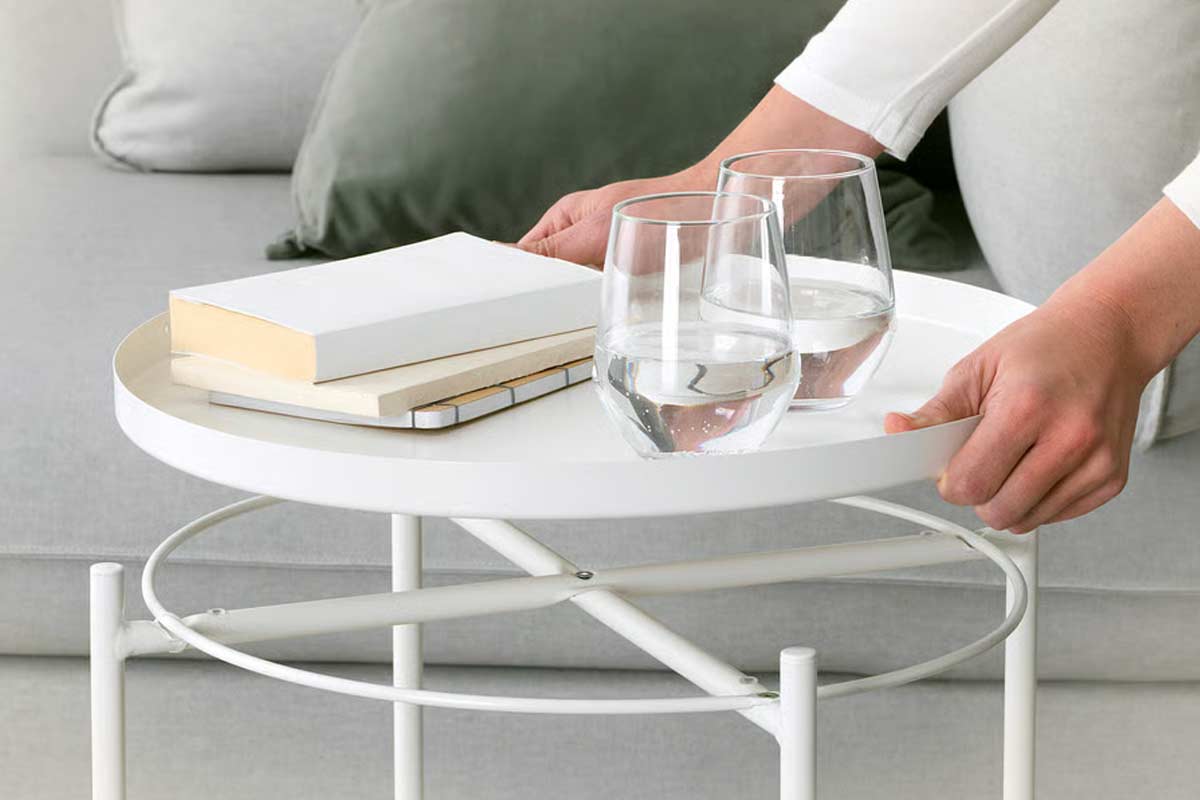 Primo piano del tavolino Gladom di IKEA in metallo bianco, con il vassoio removibile su cui sono poggiati due bicchieri d’acqua e alcuni libri, mentre una persona lo solleva con entrambe le mani davanti a un divano chiaro.