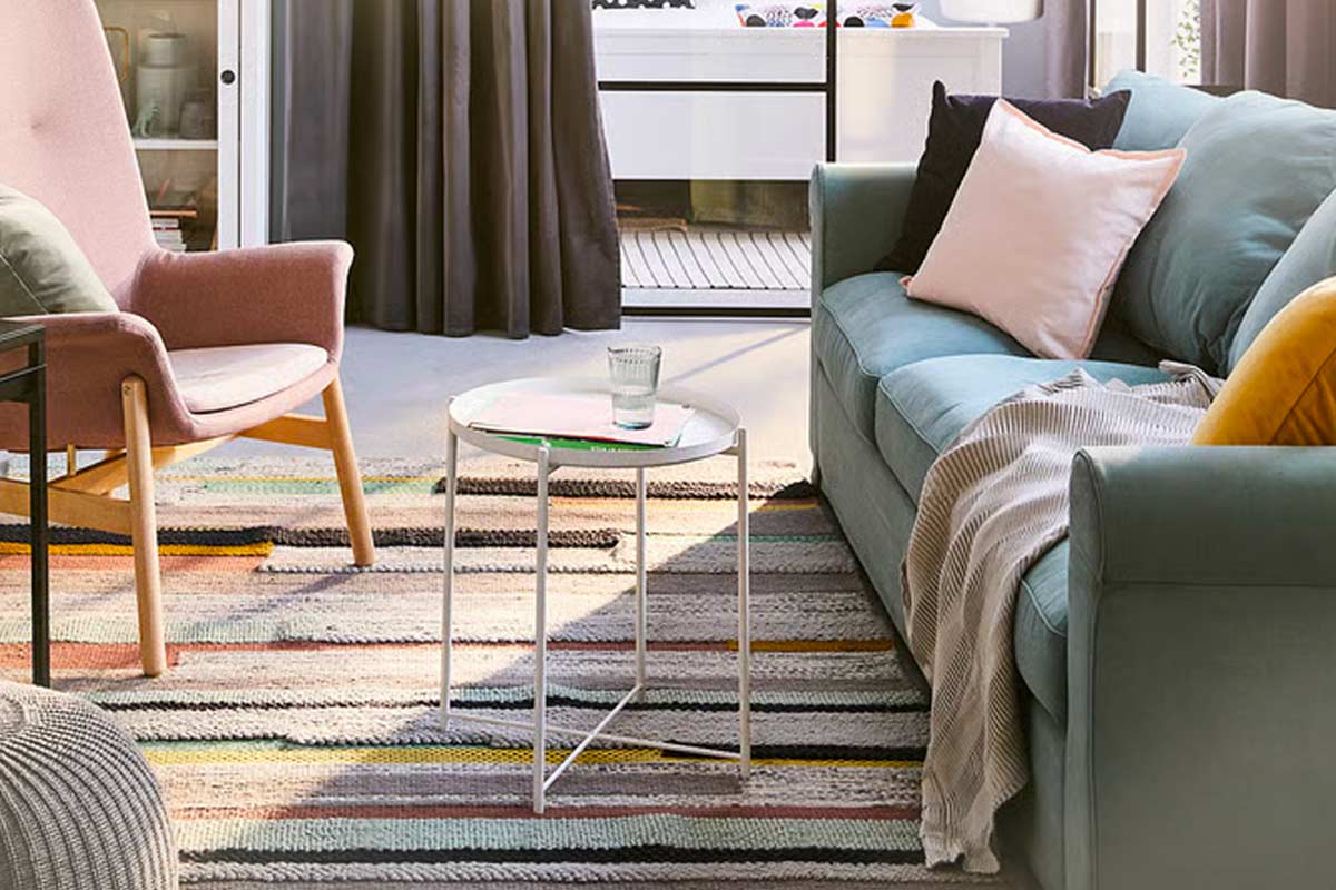 Tavolino rotondo Gladom di IKEA in metallo bianco posizionato su un tappeto colorato in un salotto moderno, accanto a un divano azzurro con cuscini e una poltrona rosa, illuminato da luce naturale proveniente da una finestra.