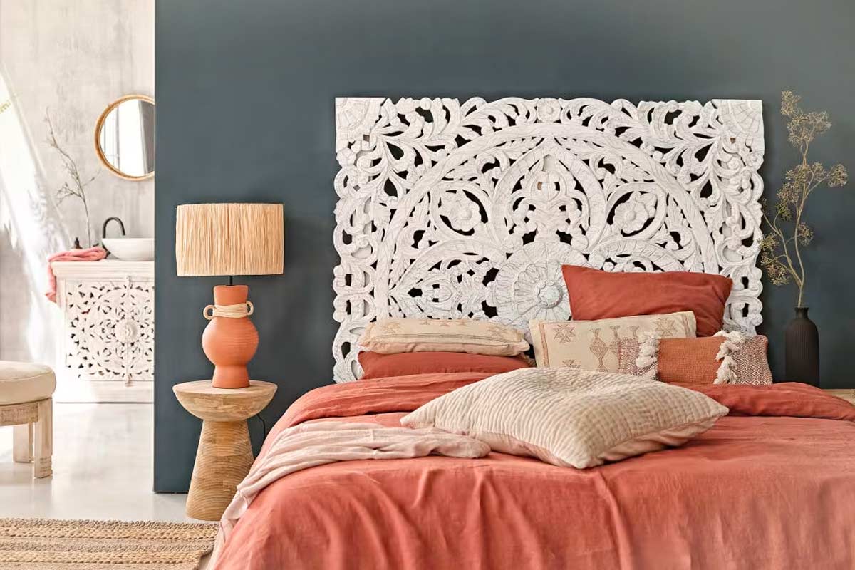 Camera da letto con parete scura, testata intagliata Kerala Maisons du Monde in legno di mango bianco, letto con biancheria e cuscini nei toni terracotta e sabbia, lampada in ceramica arancione su tavolino in legno, arredi naturali e dettagli artigianali, atmosfera boho chic luminosa e accogliente.