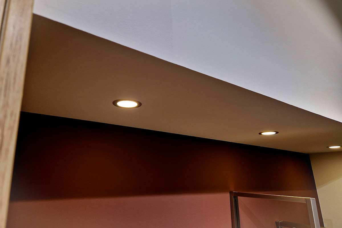 Soffitto moderno con luci incassate accese, atmosfera calda e accogliente, parete sottostante color terracotta. Linee geometriche nette e riflessi morbidi sul vetro di una porta, illuminazione soffusa.