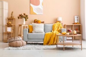 Soggiorno luminoso con pareti beige chiaro, divano grigio chiaro e tessili materici in toni neutri e giallo, tavolino in legno naturale, luce solare diffusa e decorazioni autunnali.