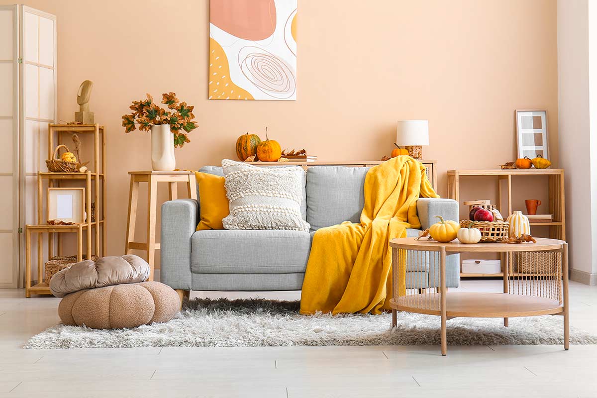 Soggiorno luminoso con pareti beige chiaro, divano grigio chiaro e tessili materici in toni neutri e giallo, tavolino in legno naturale, luce solare diffusa e decorazioni autunnali.