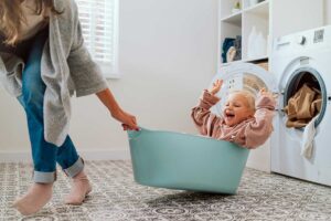 Bambino che ride mentre viene trascinato in un cesto della biancheria da un adulto, accanto a una lavatrice in una lavanderia domestica luminosa.