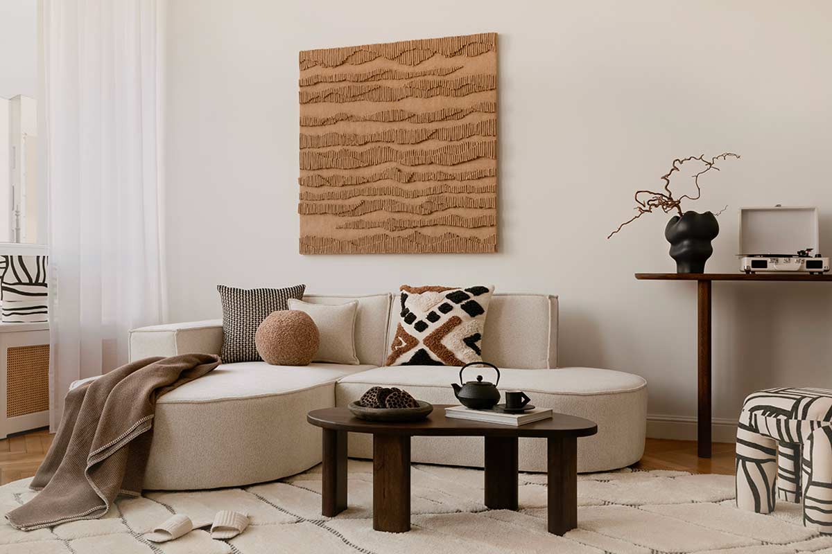 Soggiorno con parete bianco caldo, divano ton sur ton e dettagli materici, tavolino in legno scuro e cuscini grafici su tappeto chiaro.