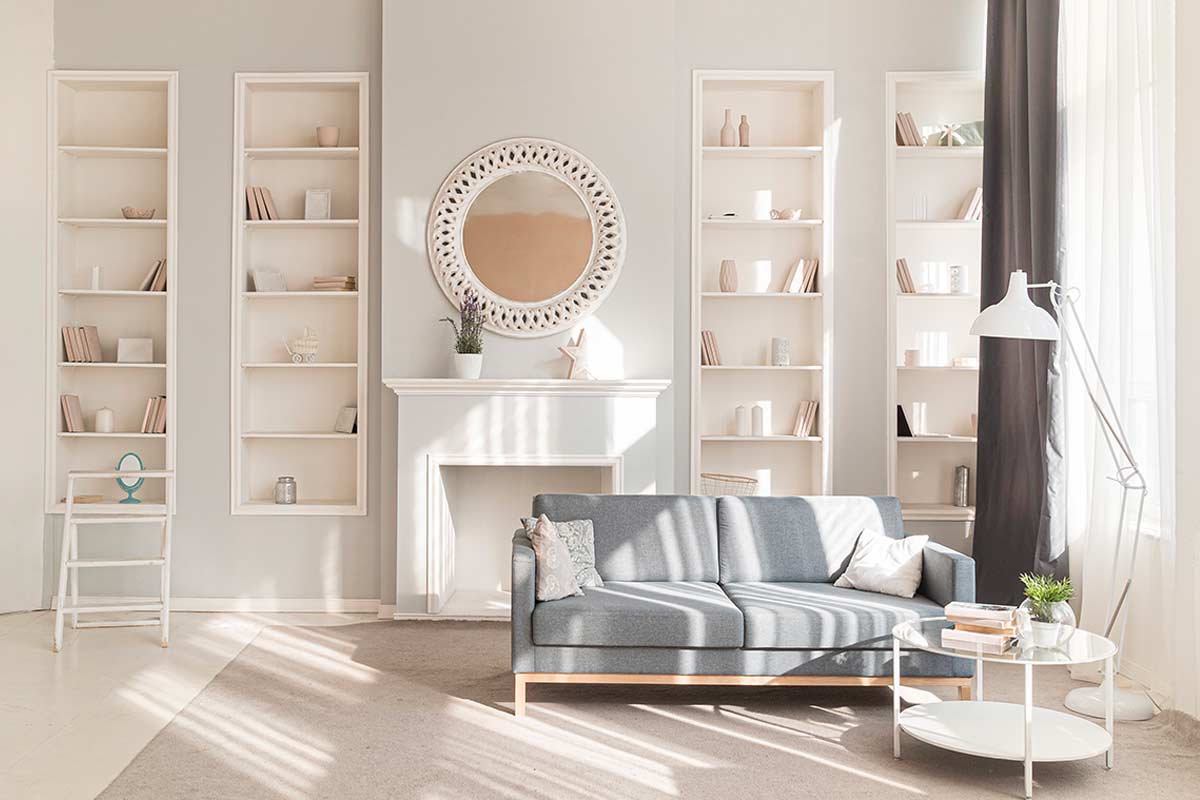 Salotto luminoso con divano grigio, specchio rotondo e librerie bianche ordinate.