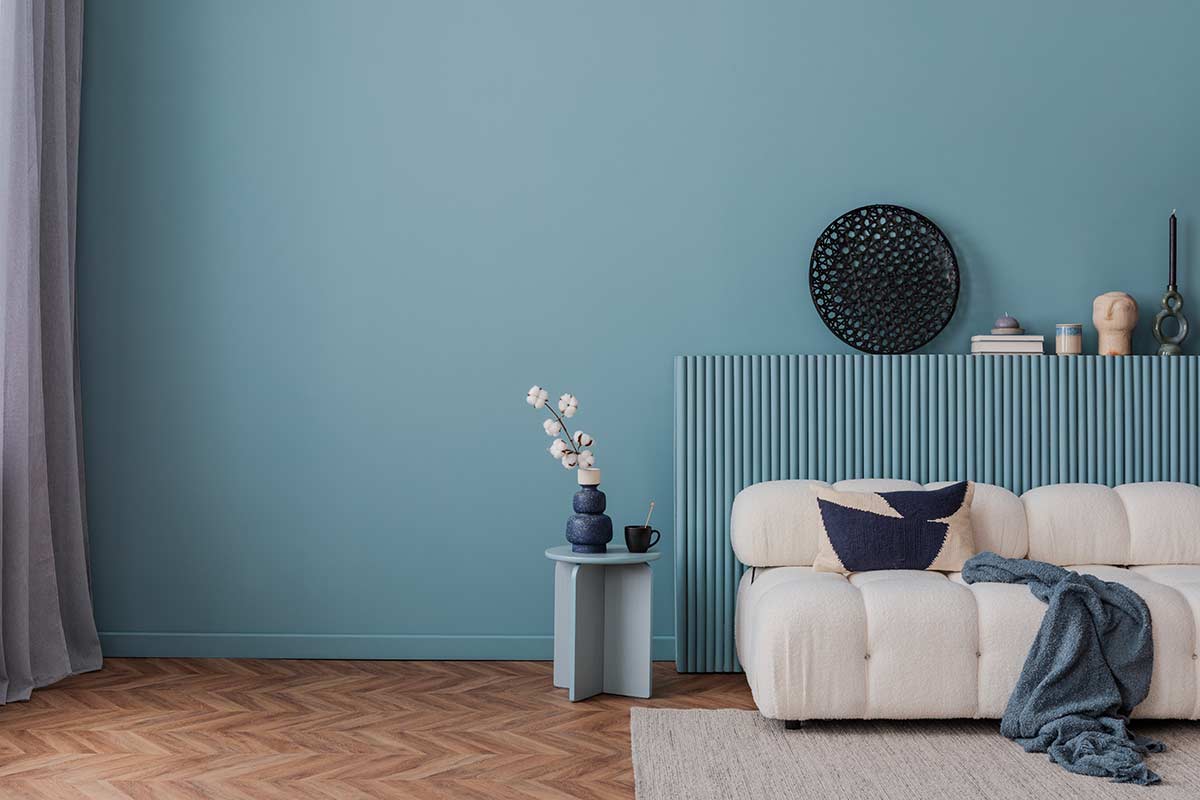 Salotto con parete blu polvere, pavimento scuro in legno, divano chiaro e dettagli minimal, luce naturale filtrata da tenda leggera.