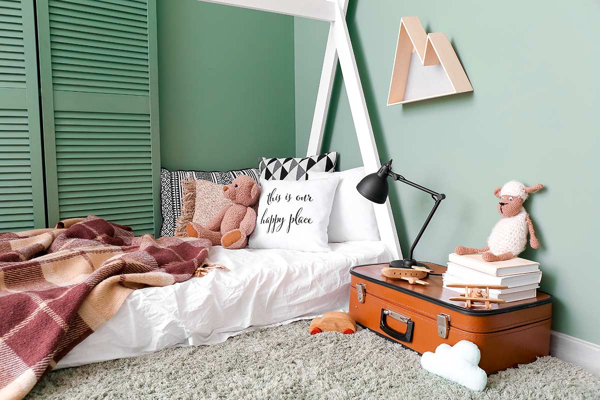 Cameretta per bambini con pareti verde salvia, letto basso con cuscini e peluche, arredi in materiali naturali e atmosfera rilassante.