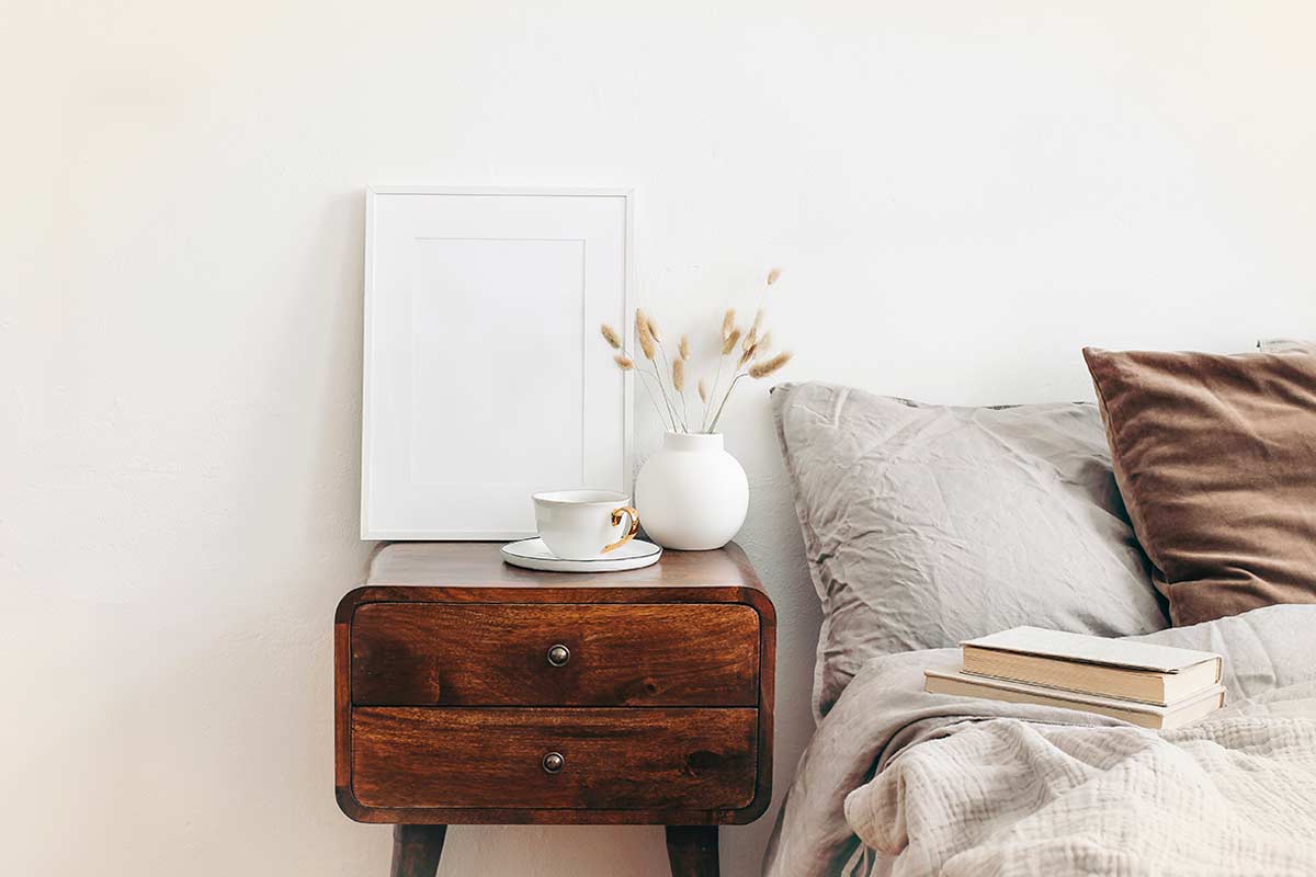 Comodino in noce anni ’50 con tazza, vaso in ceramica e letto neutro su parete chiara minimal.