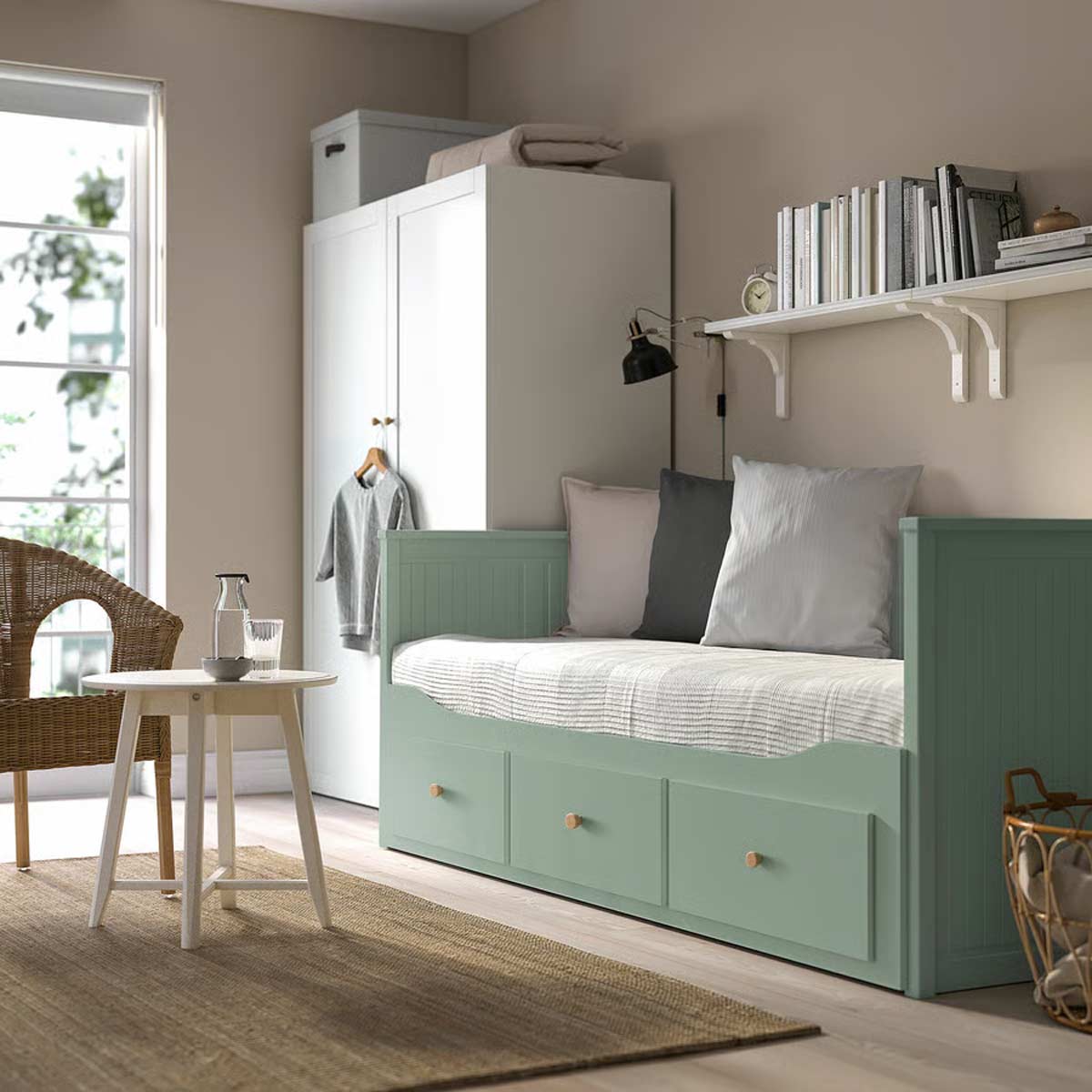 Divano letto HEMNES grigio-verde di IKEA in una stanza luminosa con pareti beige, libreria a muro e cassetti contenitori chiusi. Sul piano ci sono cuscini in tonalità neutre e tavolino rotondo bianco con bicchiere e caraffa.