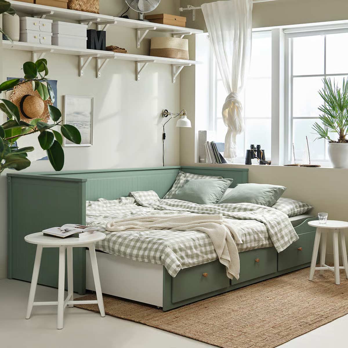 Divano letto HEMNES grigio-verde di IKEA aperto in versione matrimoniale, con biancheria a quadretti verde chiaro e cassetti contenitori. Ambiente luminoso, piante, tavolini bianchi e mensole con scatole e libri.
