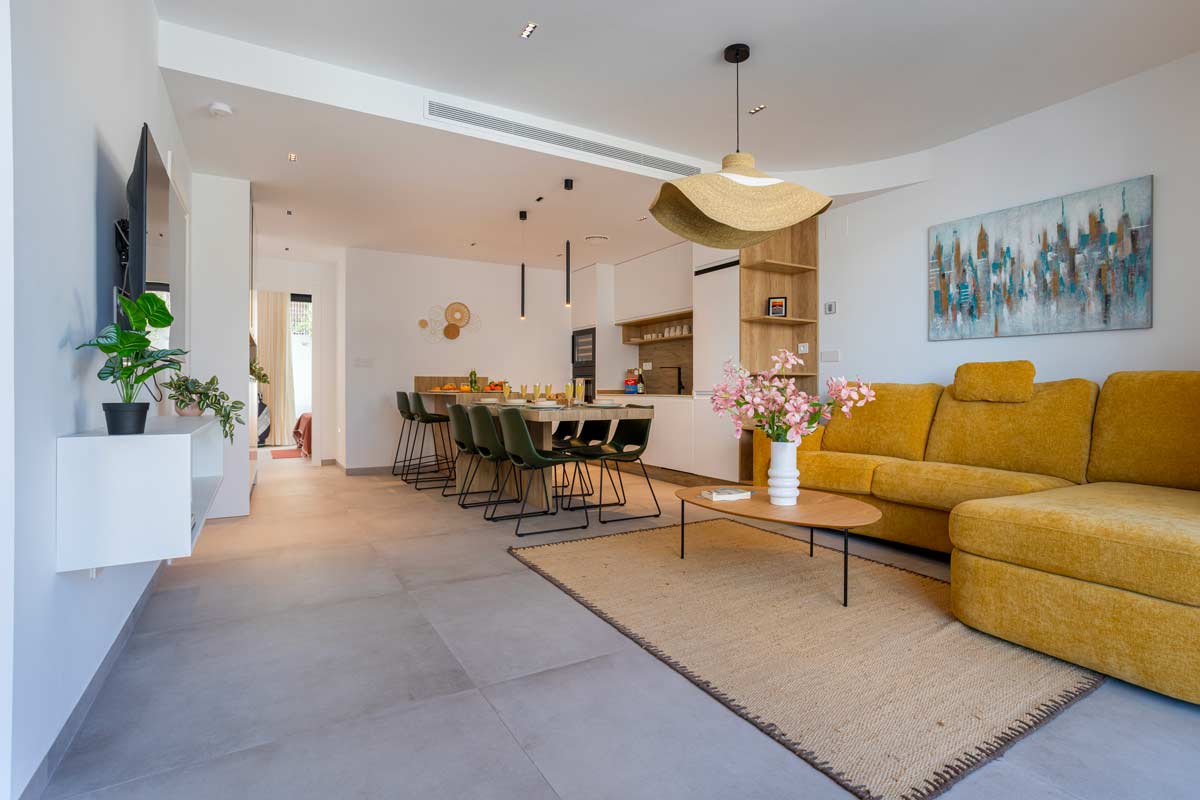 Open space contemporaneo con cucina lineare e isola centrale che introduce la zona pranzo, soggiorno con divano ocra e tappeto neutro a definire l’area relax senza separazioni murarie.