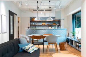 Open space moderno con penisola centrale rivestita in piastrelle azzurre che separa cucina e soggiorno, tavolo in legno e divano grigio in primo piano a definire le diverse funzioni dell’ambiente.