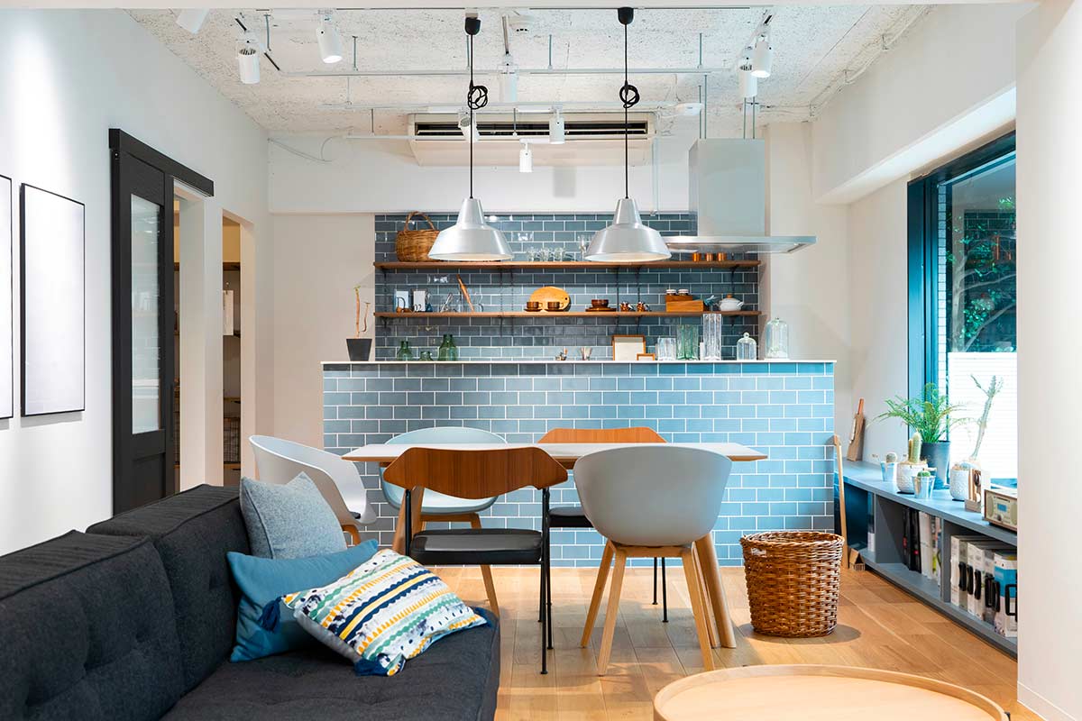 Open space moderno con penisola centrale rivestita in piastrelle azzurre che separa cucina e soggiorno, tavolo in legno e divano grigio in primo piano a definire le diverse funzioni dell’ambiente.