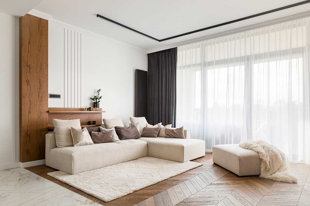 Salotto moderno e luminoso con grande divano angolare color crema, tappeto beige di misura corretta che unisce la zona conversazione, tende leggere a tutta altezza e pavimento in legno con inserti in marmo. L’arredo trasmette ordine e ampiezza grazie al tappeto posizionato secondo le regole dei designer.