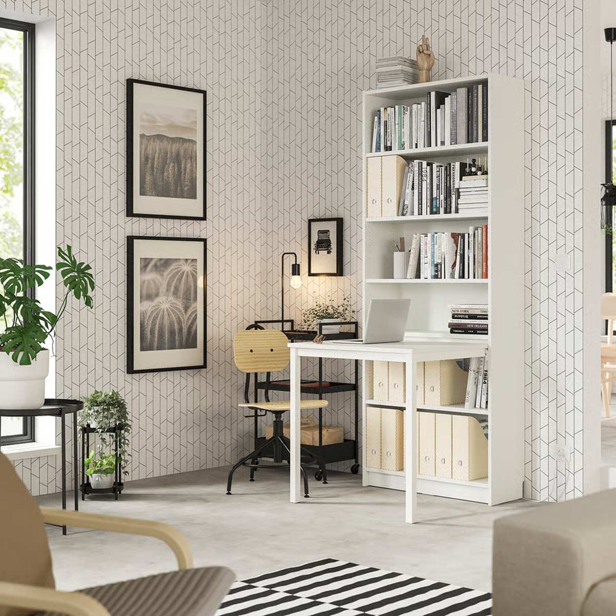 Libreria bianca BILLY di IKEA con scrivania integrata estraibile, allestita come postazione smart working in un soggiorno moderno, con scatole archivio, libri e laptop, su sfondo di carta da parati geometrica chiara.