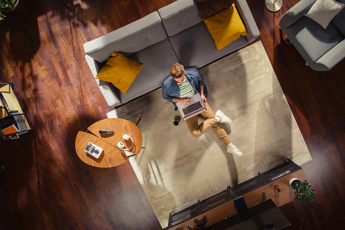 Vista dall’alto di un soggiorno moderno con divano grigio staccato dalla parete, cuscini gialli, tappeto chiaro, tavolino rotondo in legno e una persona seduta con un laptop.