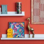 Mensole bianche su parete arancione con oggetti decorativi disposti a gruppi, spazio vuoto e dettagli colorati.
