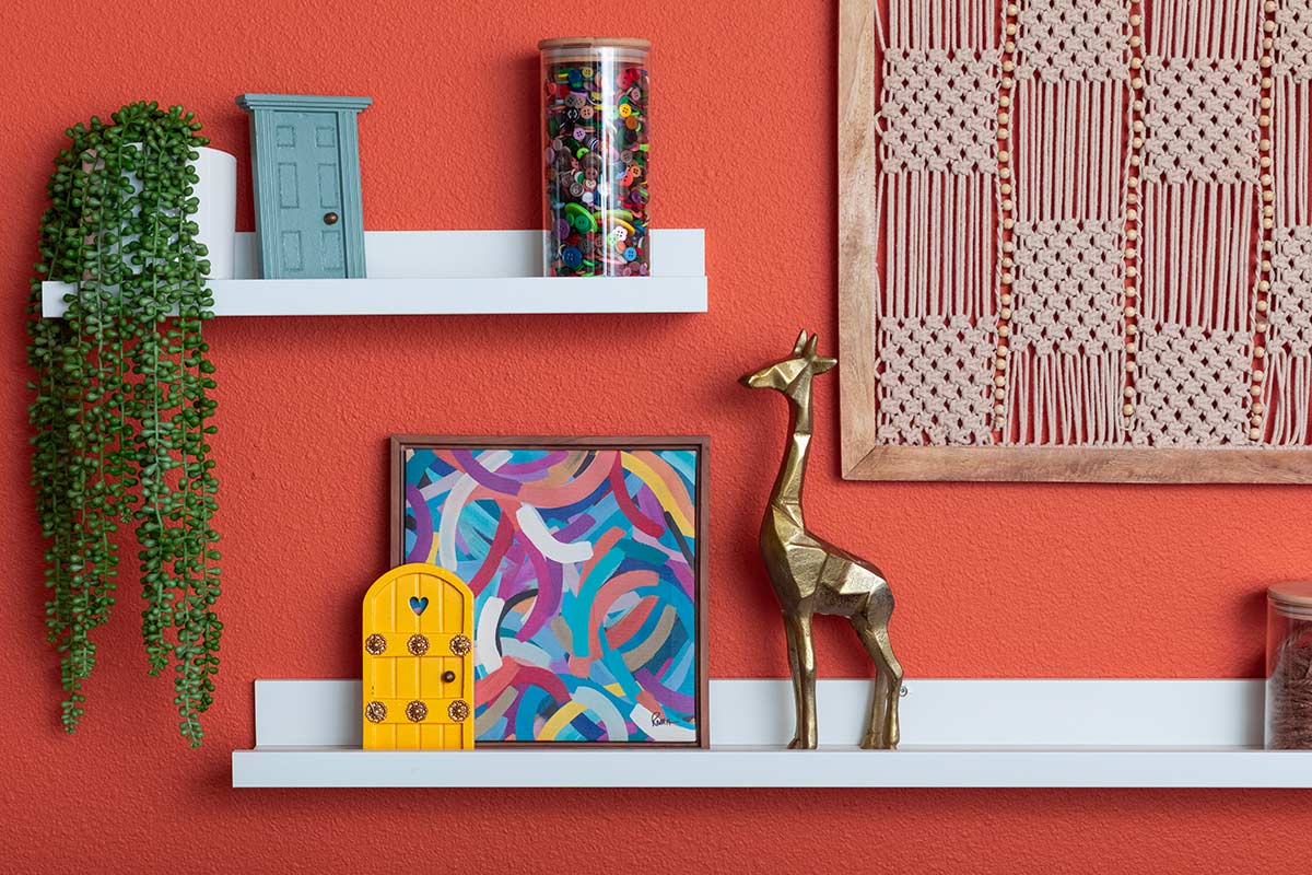 Mensole bianche su parete arancione con oggetti decorativi disposti a gruppi, spazio vuoto e dettagli colorati.
