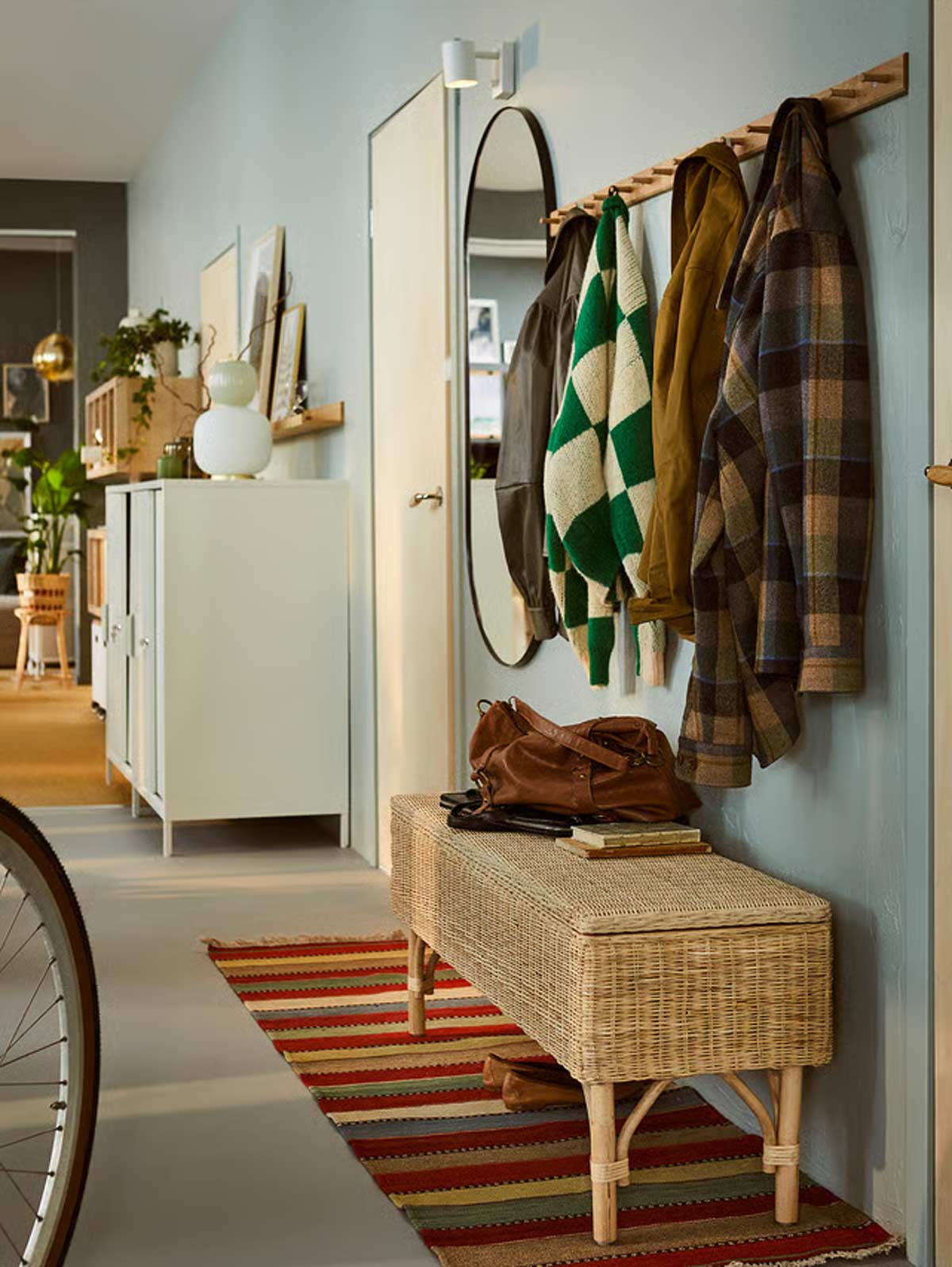 Ingresso arredato con panca contenitore IKEA TOLKNING in rattan, tappeto a righe e cappotti appesi alla parete.