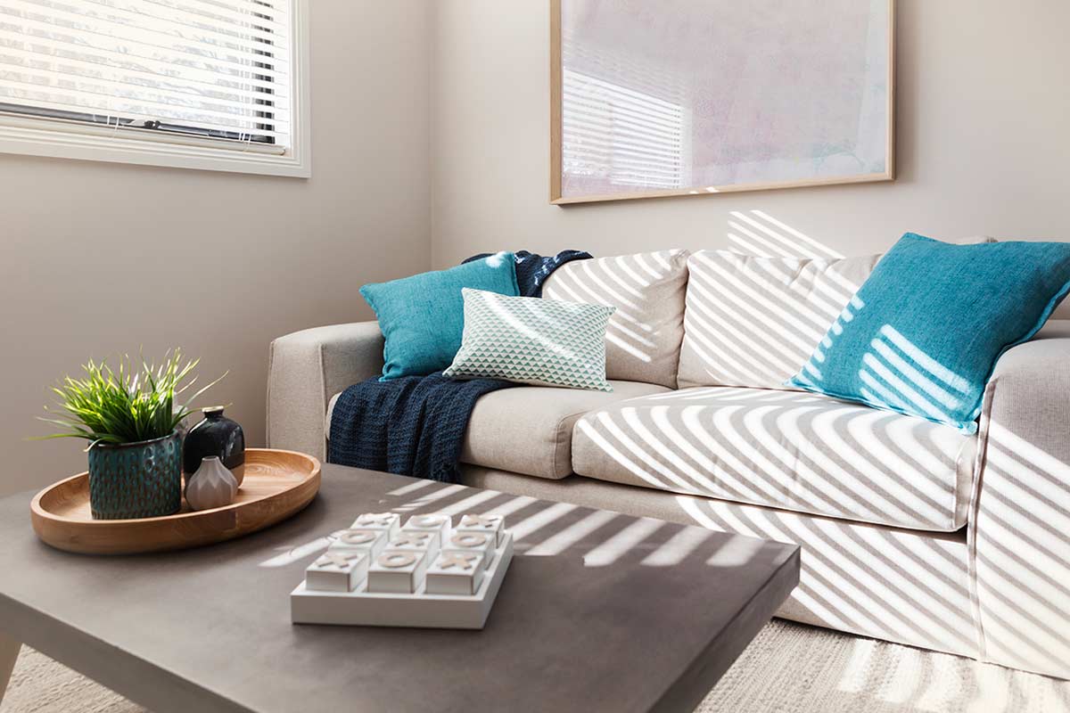 Soggiorno con pareti tortora chiare, divano beige con cuscini turchesi e tavolino moderno in primo piano illuminato da luce naturale filtrata.