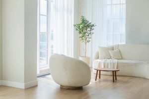 Soggiorno luminoso con ampio spazio vuoto, poltrona e tavolino distanziati dal divano, pianta in angolo e luce naturale su pavimento in legno.