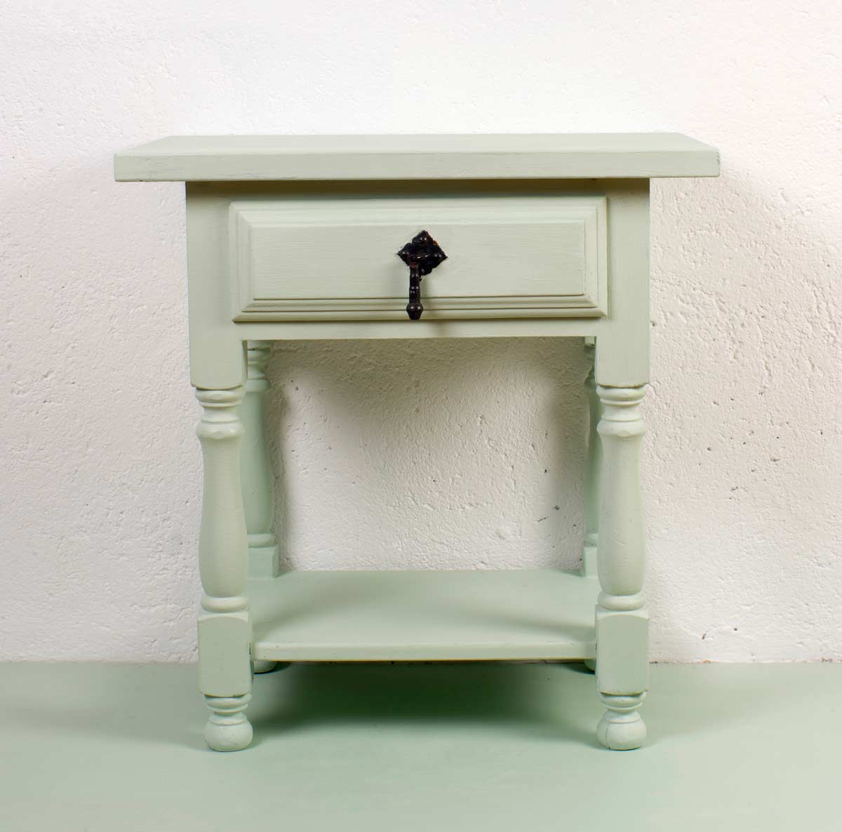 Comodino antico restaurato e verniciato in verde salvia, con gambe tornite e maniglia nera in ferro battuto, su sfondo e pavimento coordinati.