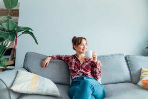Donna rilassata seduta su un divano grigio con una tazza in mano, in un soggiorno ordinato e luminoso con piante verdi e cuscini decorativi.