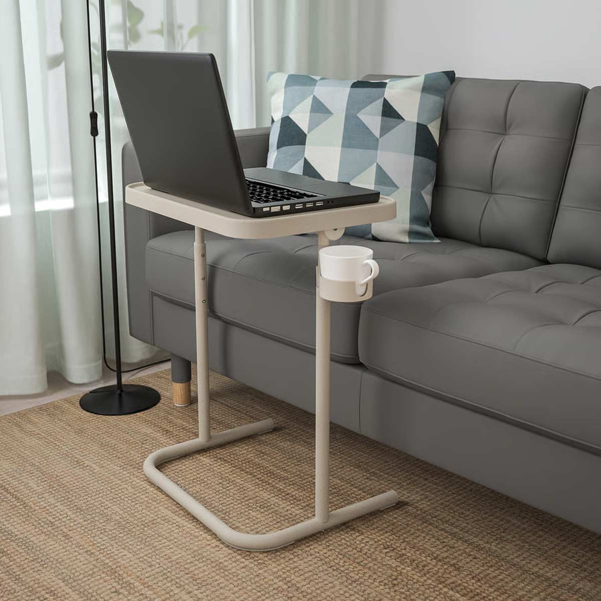 Tavolino BJÖRKÅSEN beige di IKEA con supporto per PC portatile e portatazze integrato, posizionato accanto a un divano grigio in un soggiorno moderno.