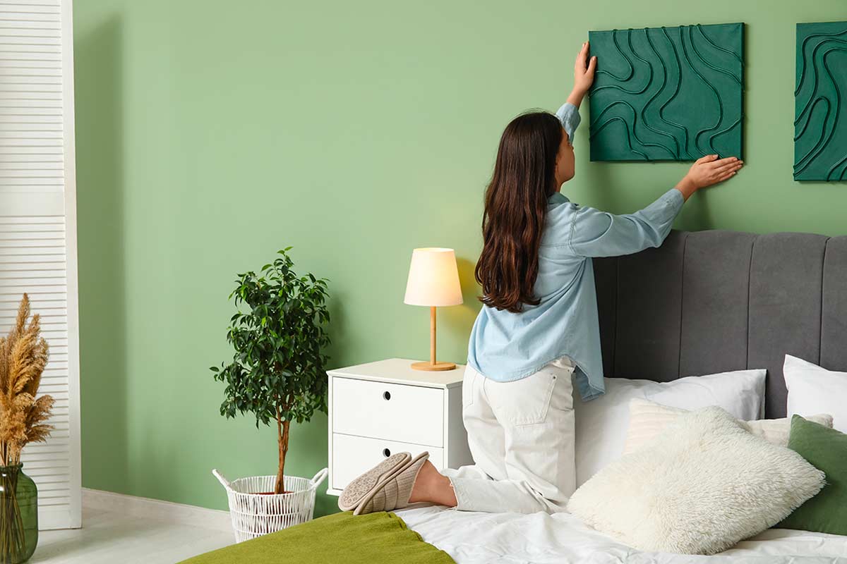 Donna che sistema un quadro su una parete verde in camera da letto moderna, con letto ordinato, lampada da comodino accesa e pianta decorativa in un angolo.