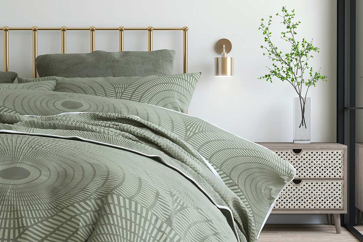 Dettaglio di camera da letto con testiera in metallo dorato, biancheria verde salvia a motivo geometrico, comodino in legno con cassetti in paglia di Vienna e vaso con rami verdi.