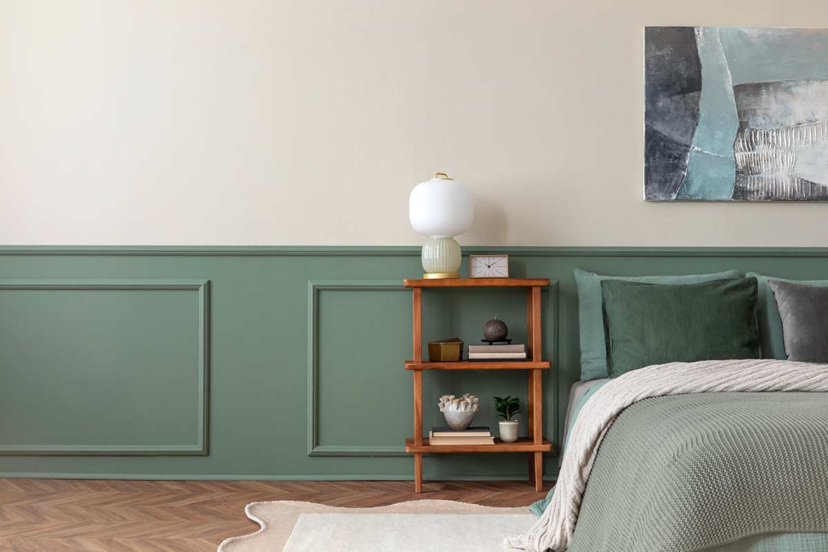 Camera da letto elegante con parete in boiserie verde salvia, letto ordinato con cuscini tono su tono, comodino in legno e lampada da tavolo, quadro astratto appeso alla parete.