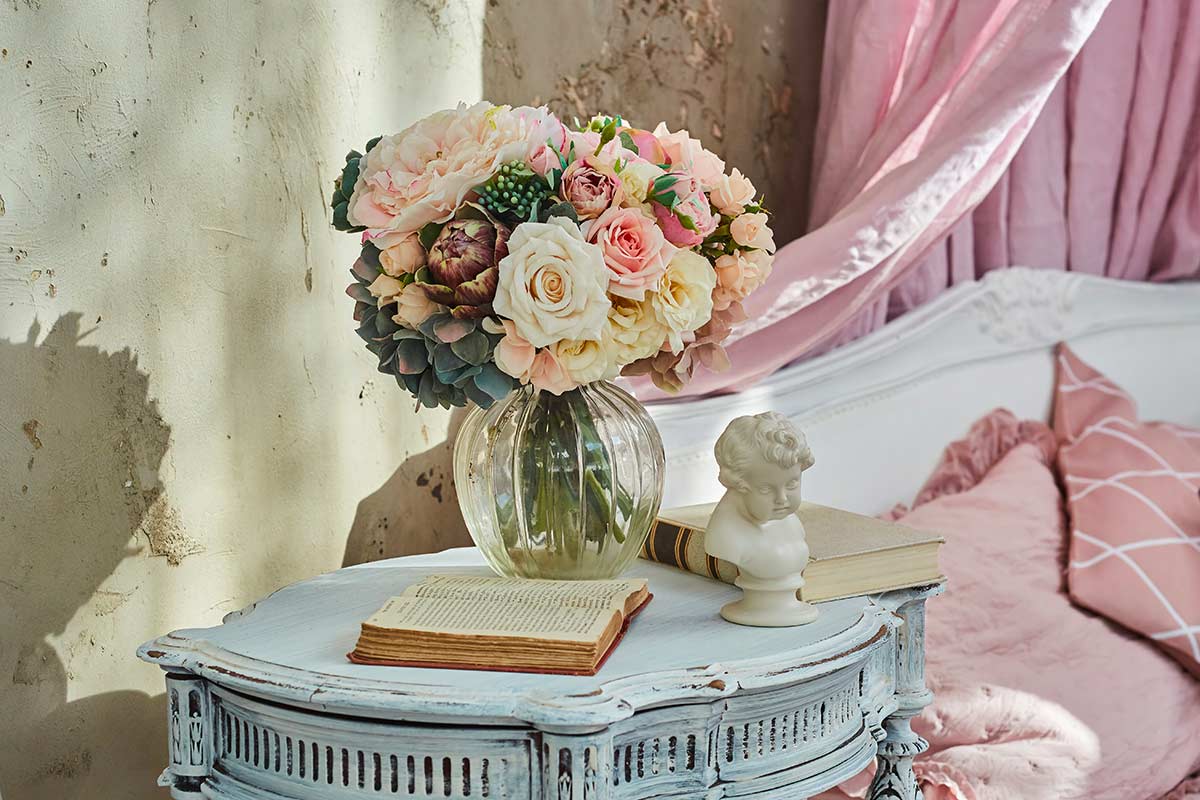Composizione floreale dai toni pastello in vaso di vetro su tavolino decapato azzurro, con libri antichi e dettagli romantici in stile Shabby Chic.