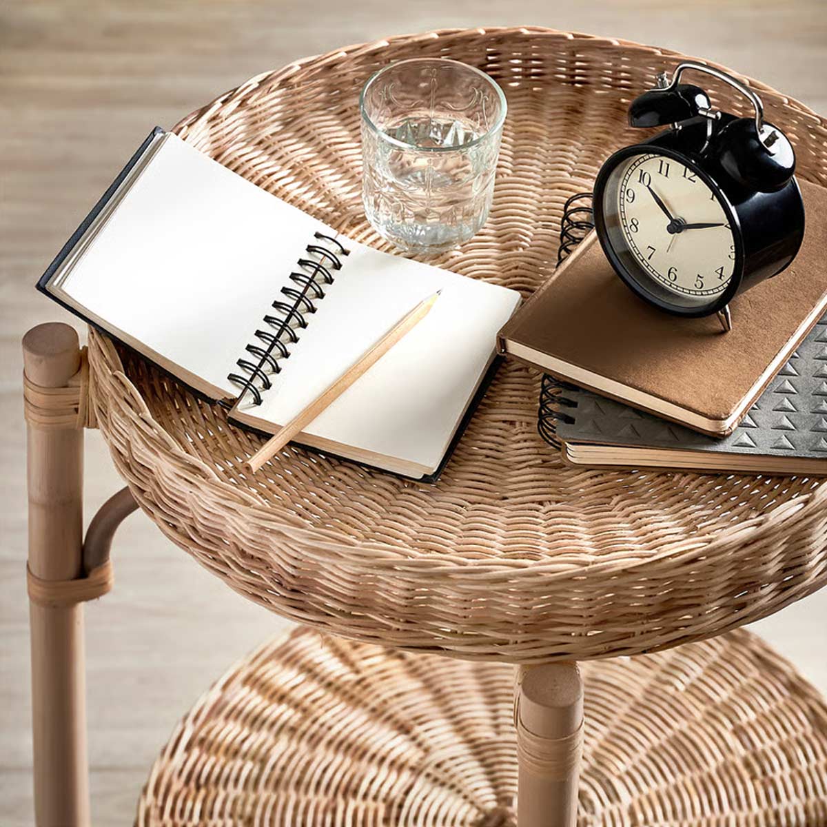 Comodino IKEA in rattan con vassoio intrecciato, blocco note aperto, bicchiere d’acqua e sveglia analogica.