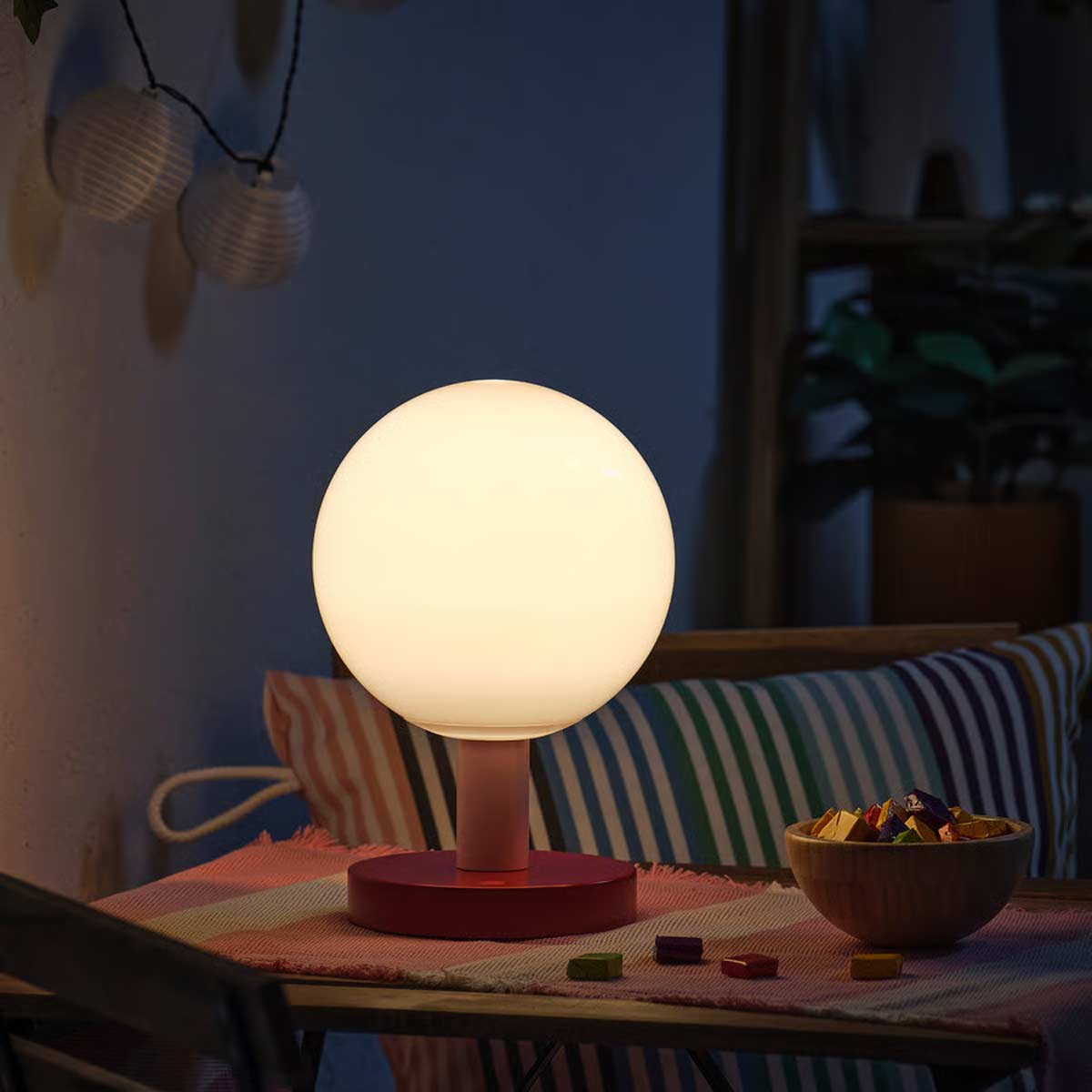 Lampada a globo IKEA accesa su tavolo all’aperto, base rossa e sfondo con cuscini colorati.
