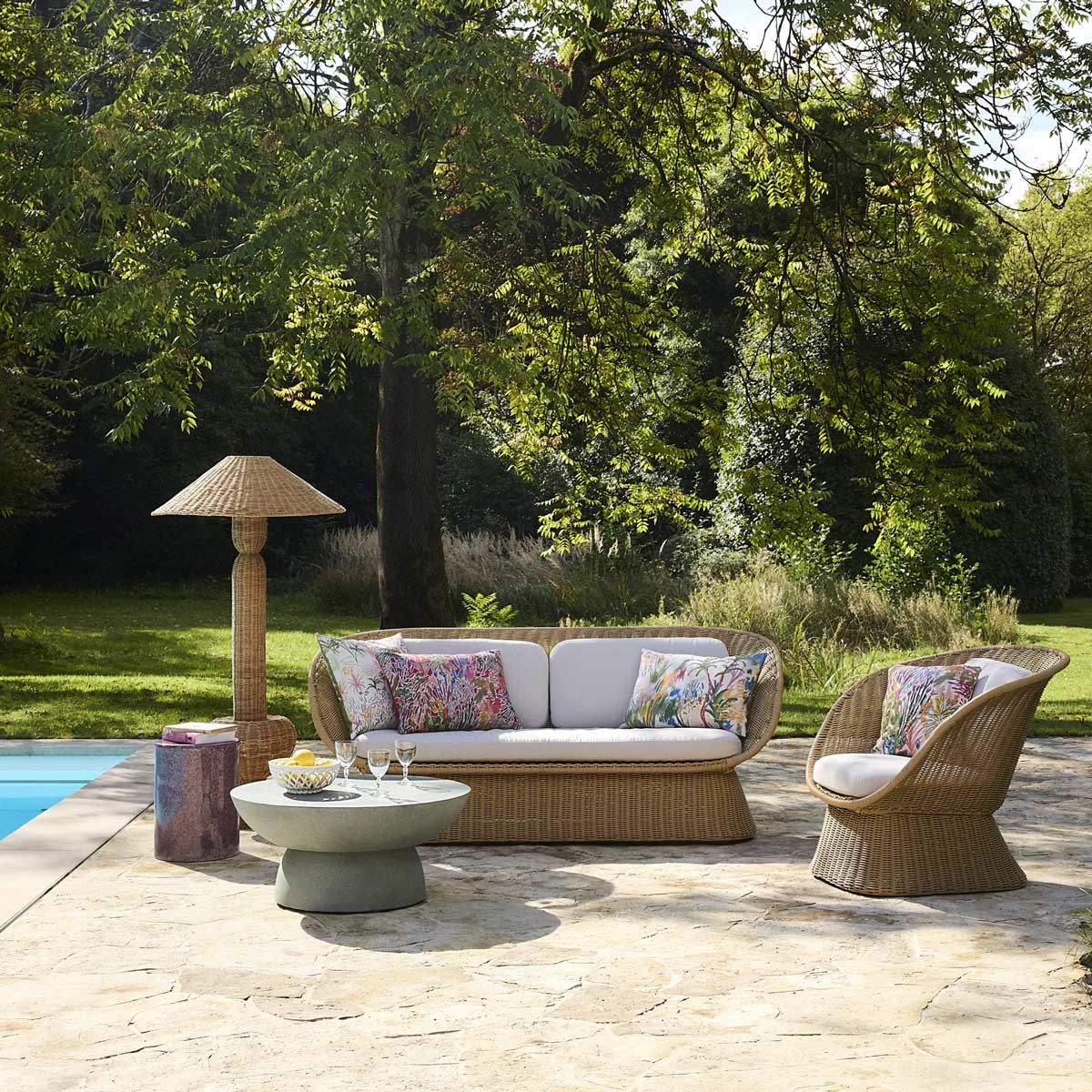 Lampada outdoor in rattan accanto a divano e poltrona da giardino su terrazza con piscina, ambiente lounge elegante immerso nel verde