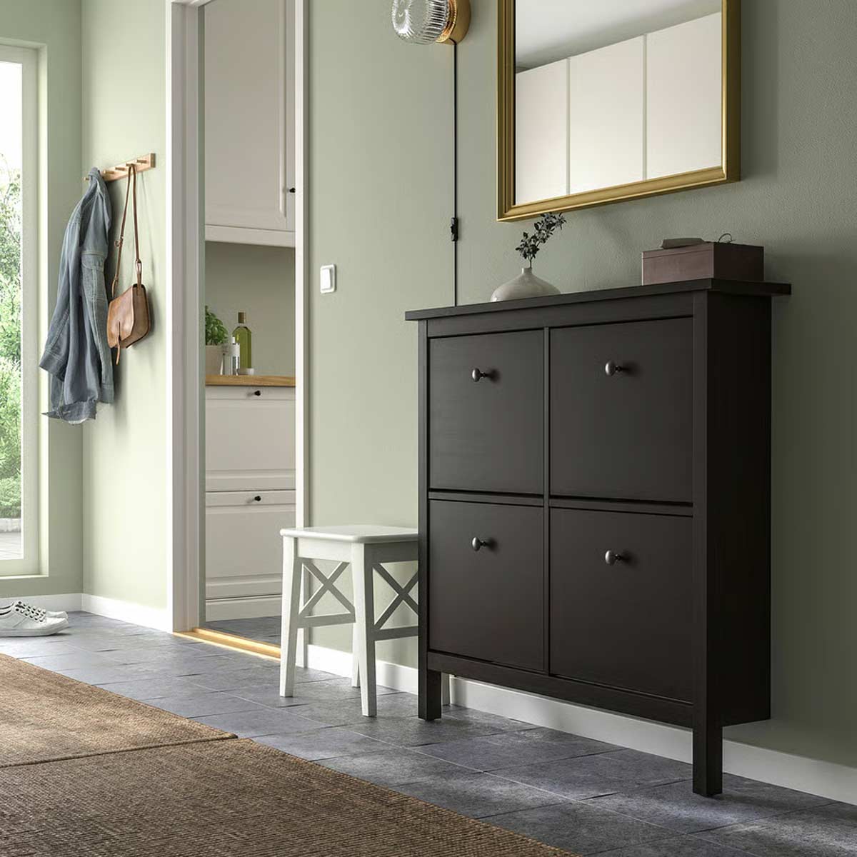 Ingresso moderno con scarpiera HEMNES marrone-nero IKEA, specchio dorato, sgabello bianco e pareti verde chiaro.