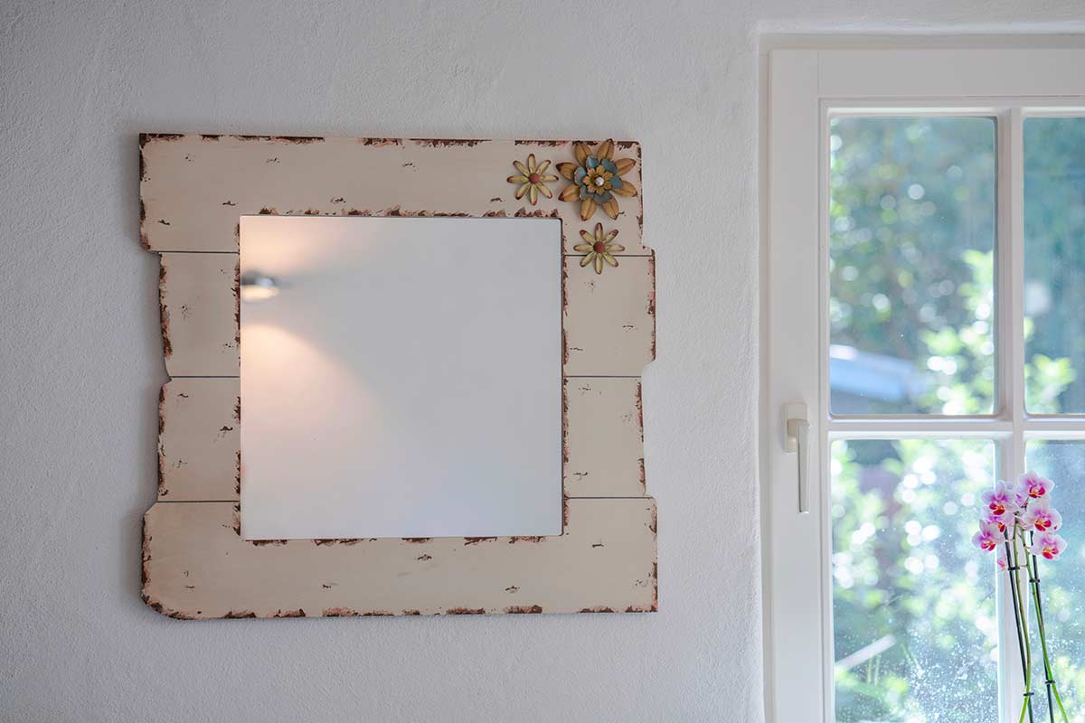 Specchio shabby chic quadrato con cornice in legno decapato color crema e decorazioni floreali applicate, montato su parete chiara accanto a una finestra con luce naturale.